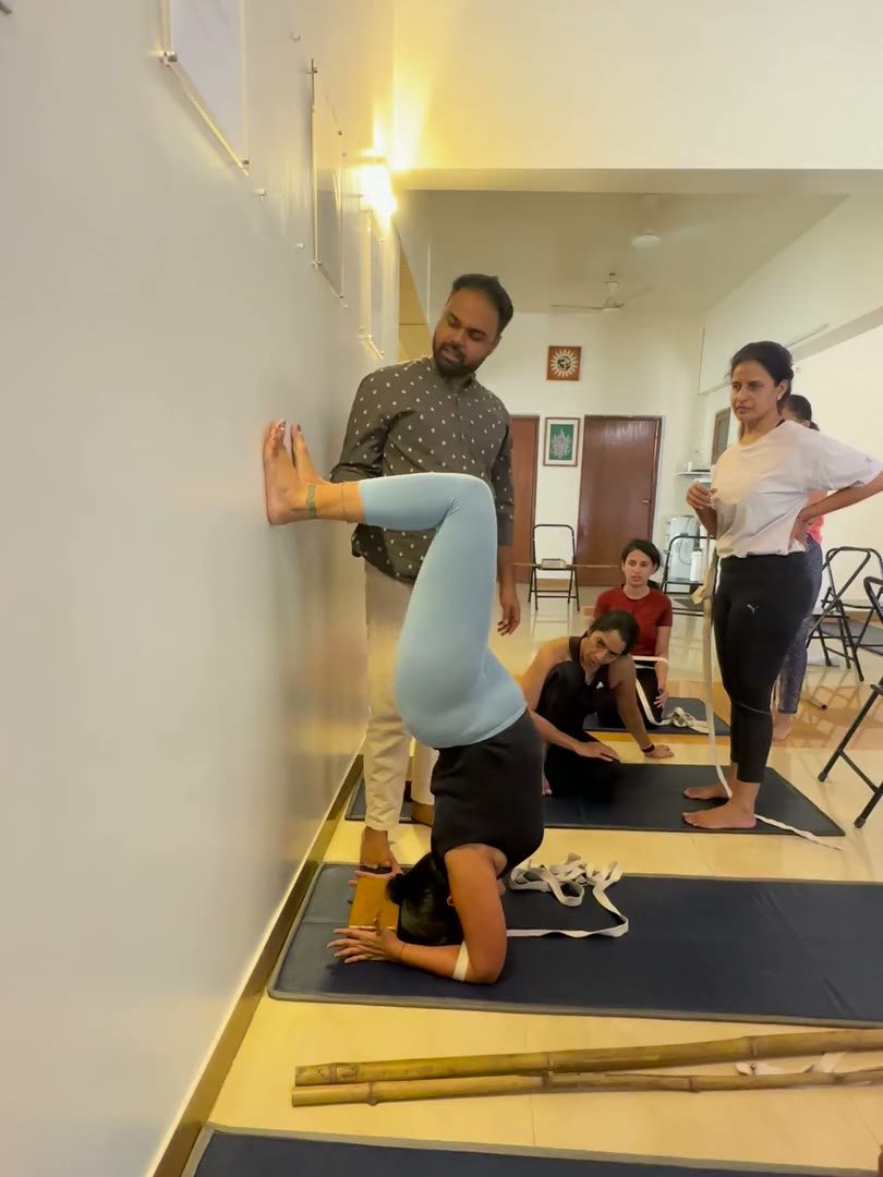 Praṇav Śāstrī beside a student balancing on her forearms, her legs arched over to the wall