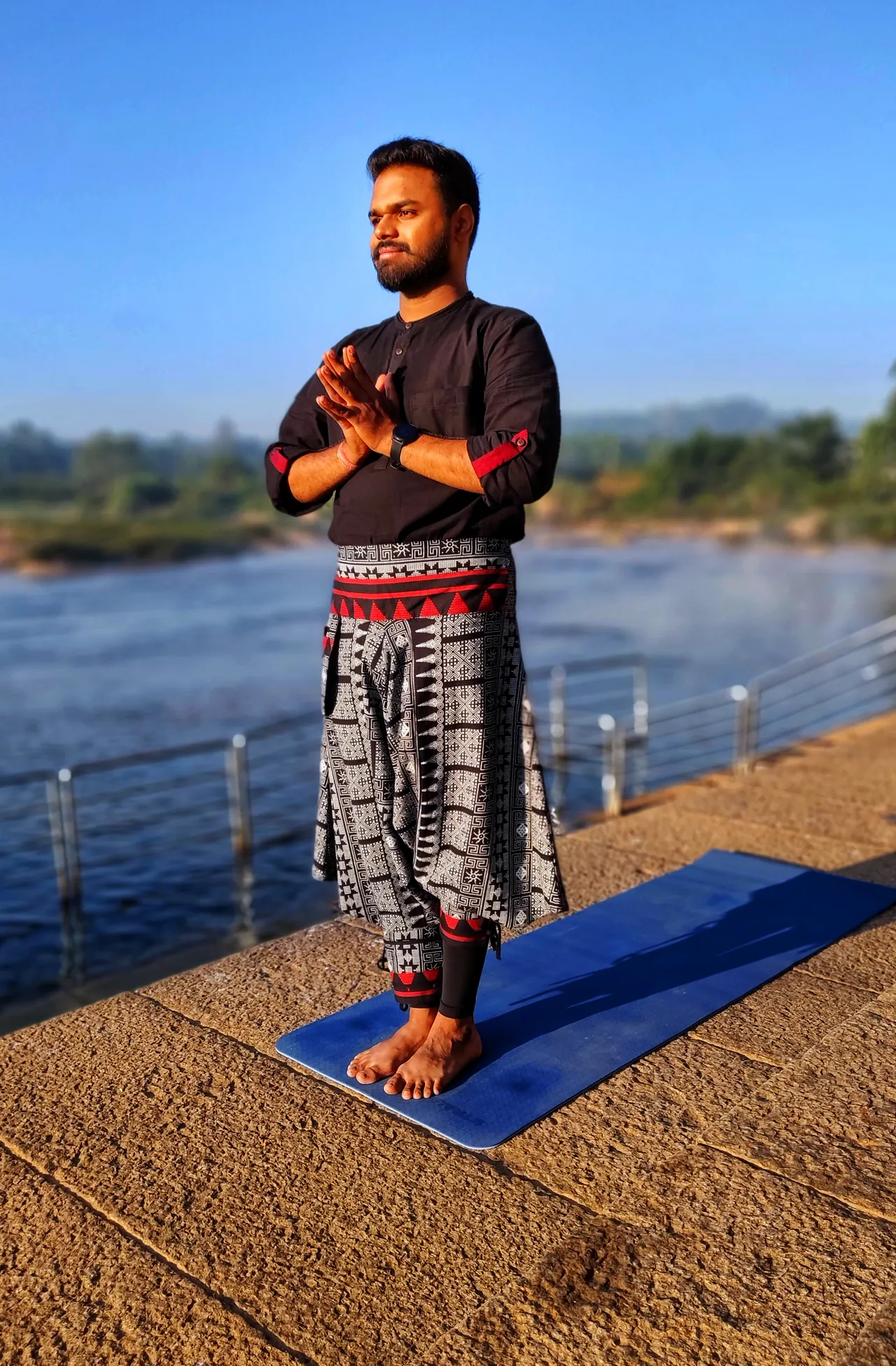 Praṇav Śāstrī standing with palms joined on a mat by a river at sunrise