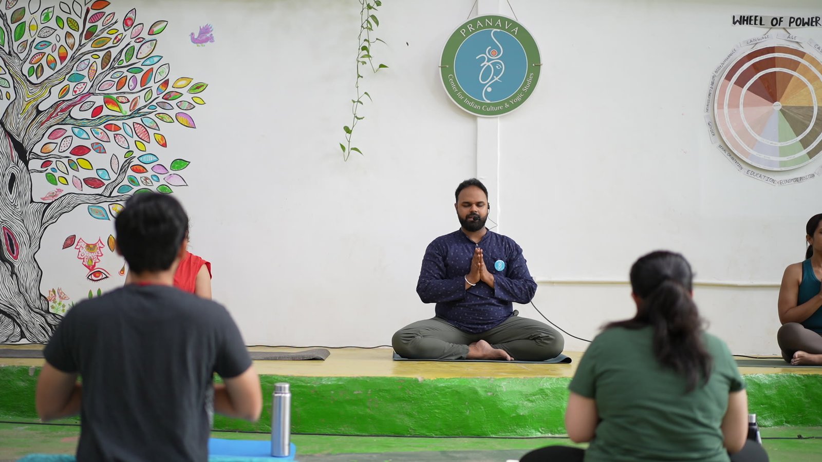 Praṇav Śāstrī seated with palms joined beneath the Praṇava emblem, his class seated in front of him