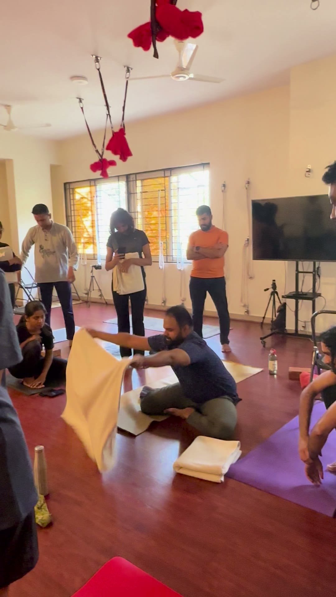 A man kneels spreading a blanket over a mat while a woman crouches beside him and three people stand talking behind them in the studio.