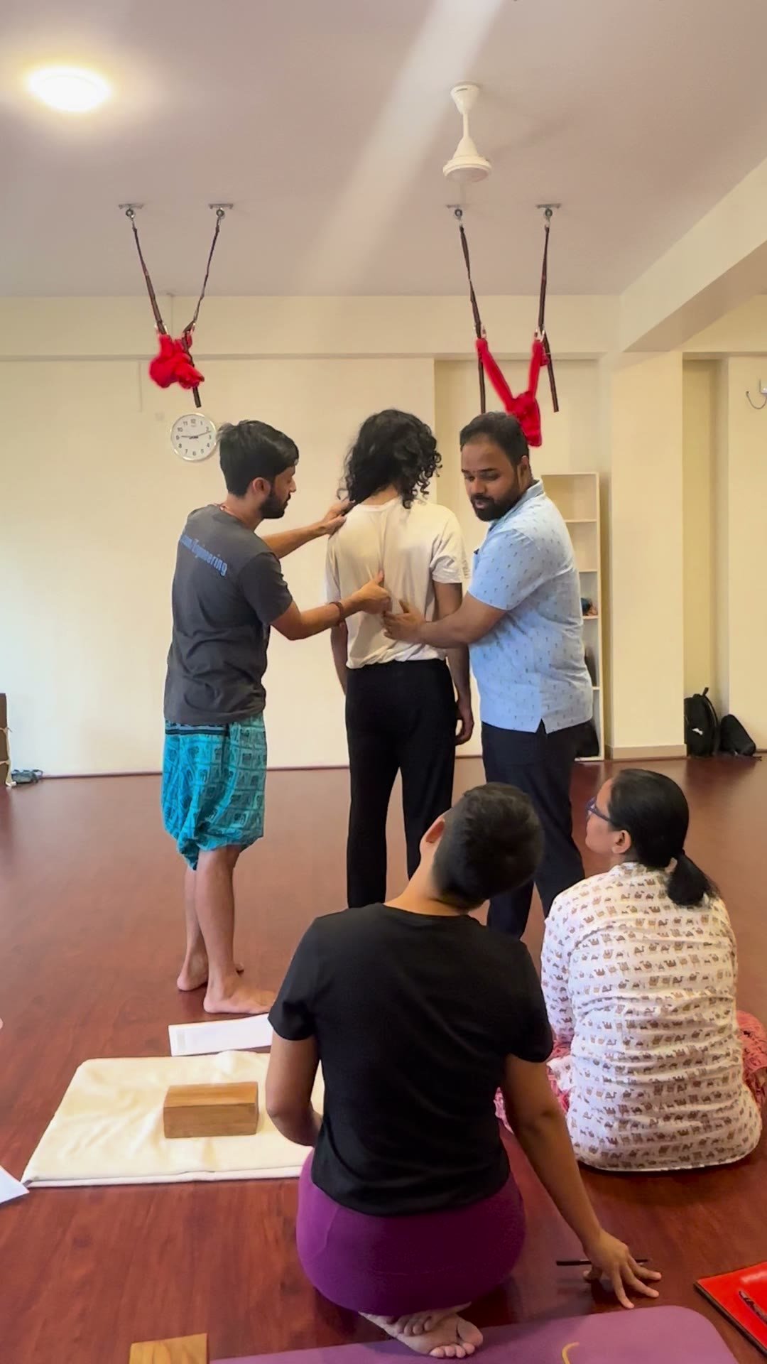 Two people work together on a third, one with a hand at her shoulders and the other at her mid-back, while two more watch from the floor.