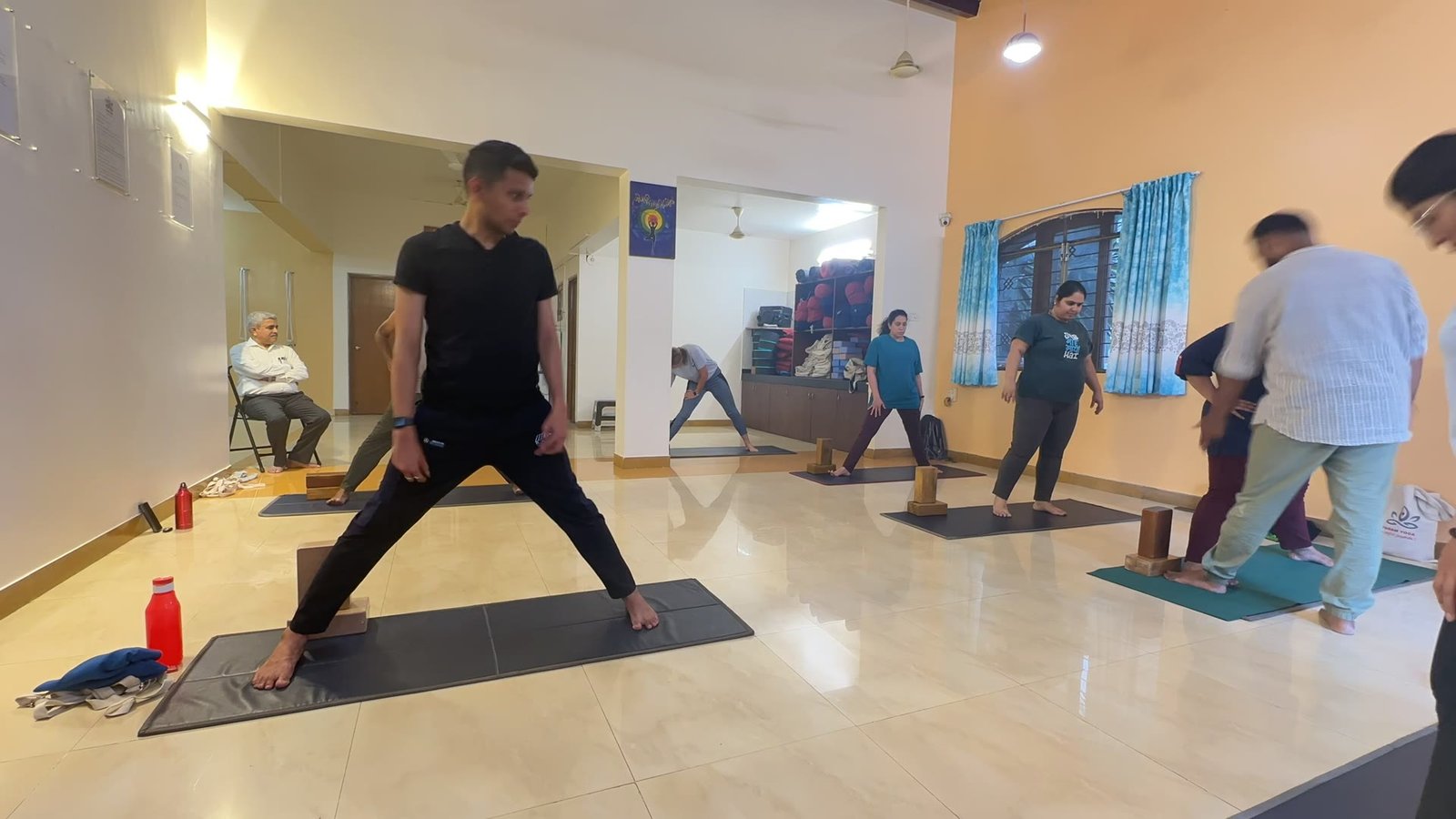 A class moving through a wide standing pose with arms extended on mats across a hall
