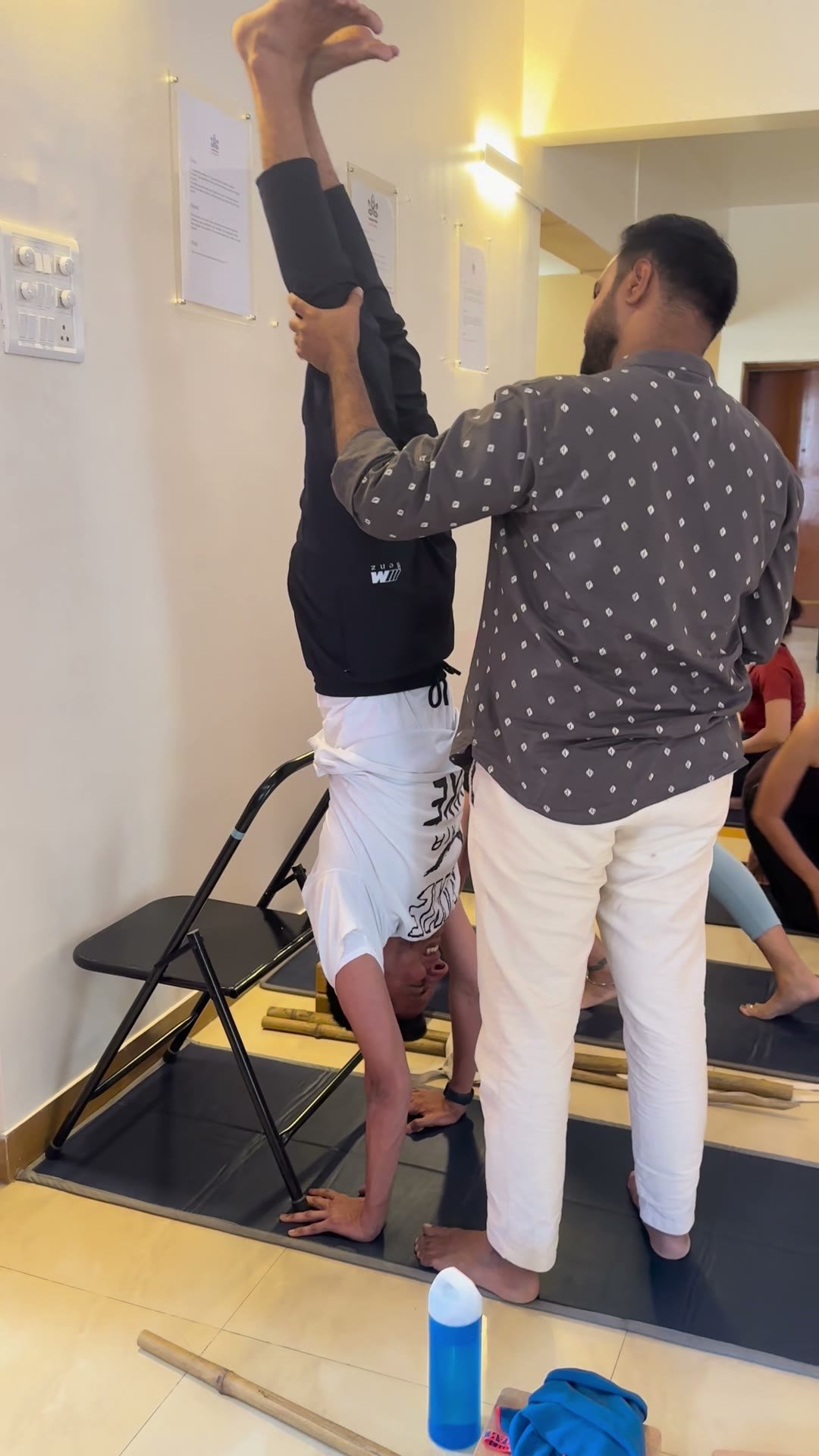 A man steadying a person holding an inverted position with their back to a wall, legs vertical