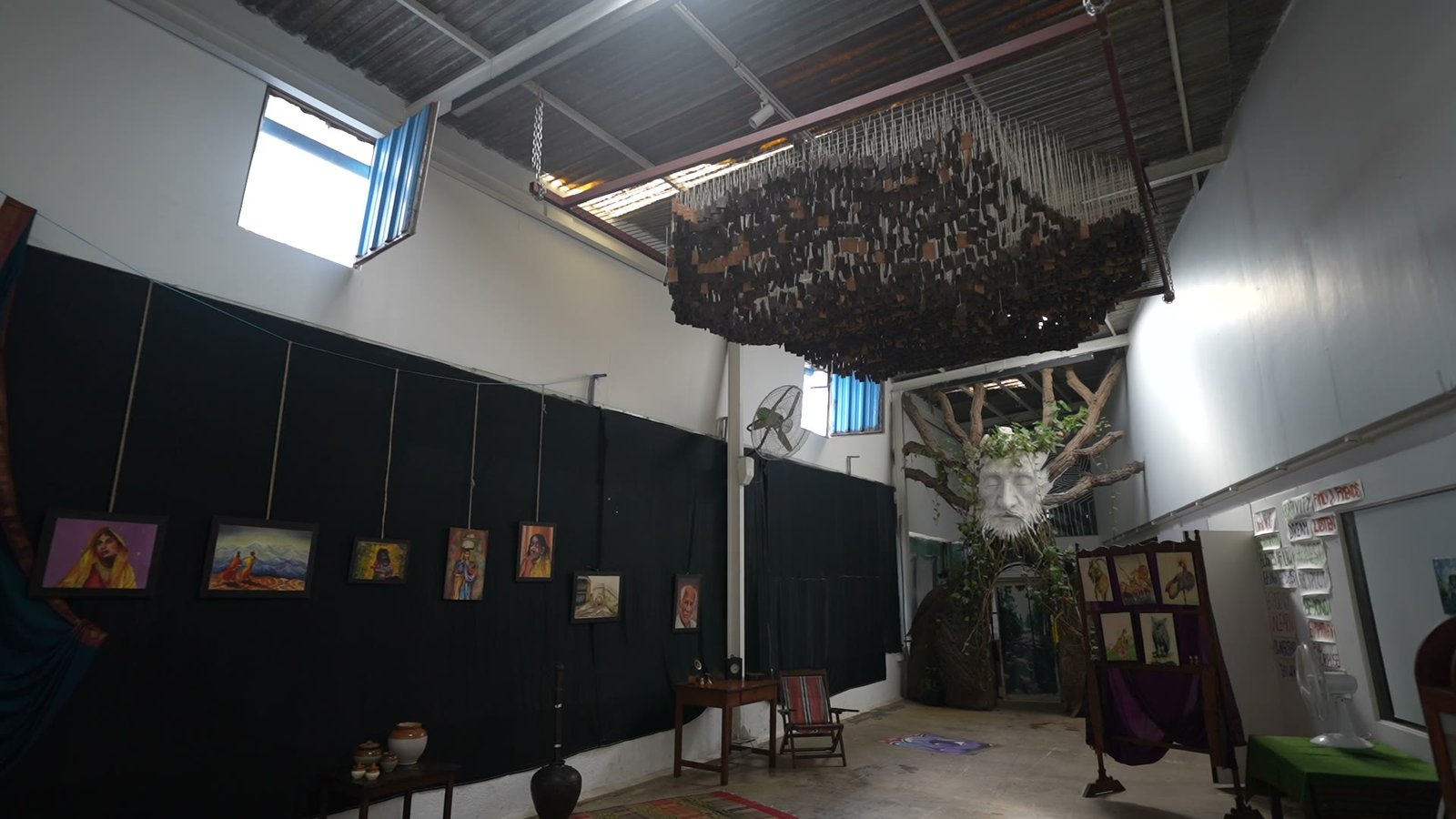 An empty exhibition hall with framed paintings hung along a dark wall, a hanging installation of wooden blocks overhead and a moulded face in a tree at the far end.