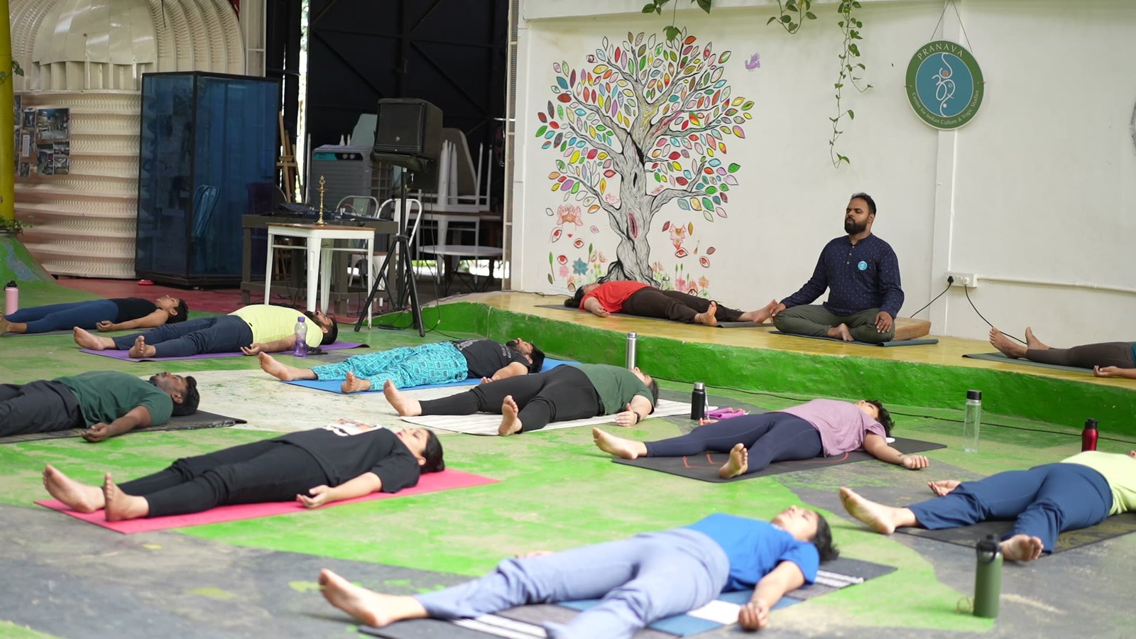 Students lying still on their mats at the end of practice, the teacher keeping watch beside them.