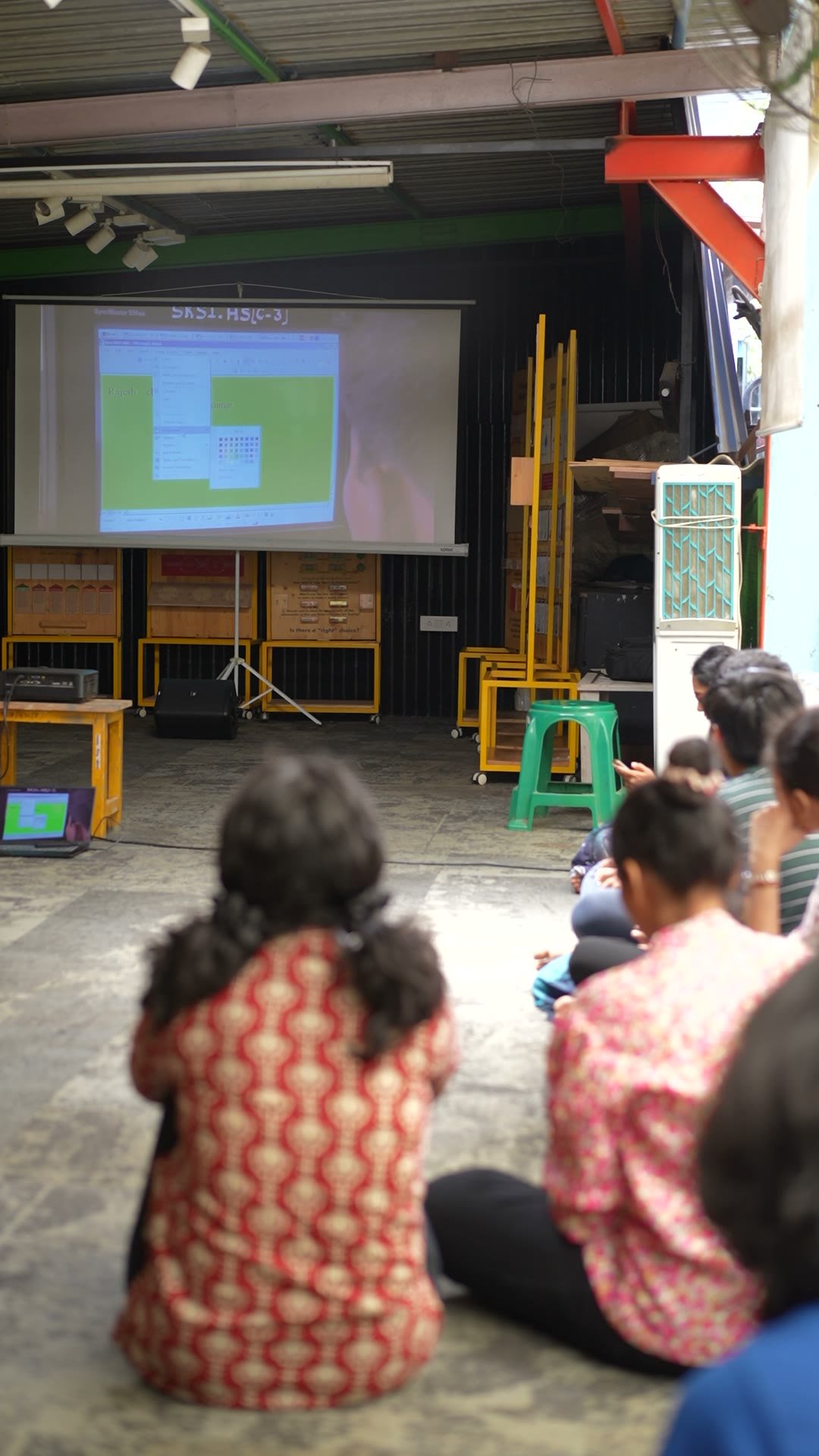 Seen from behind a seated audience, a lit projection screen at the far end of a covered yard.