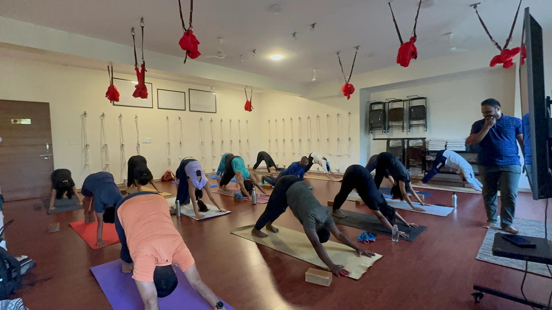 A full class holding downward-facing dog on mats while a teacher watches from the side of the hall.