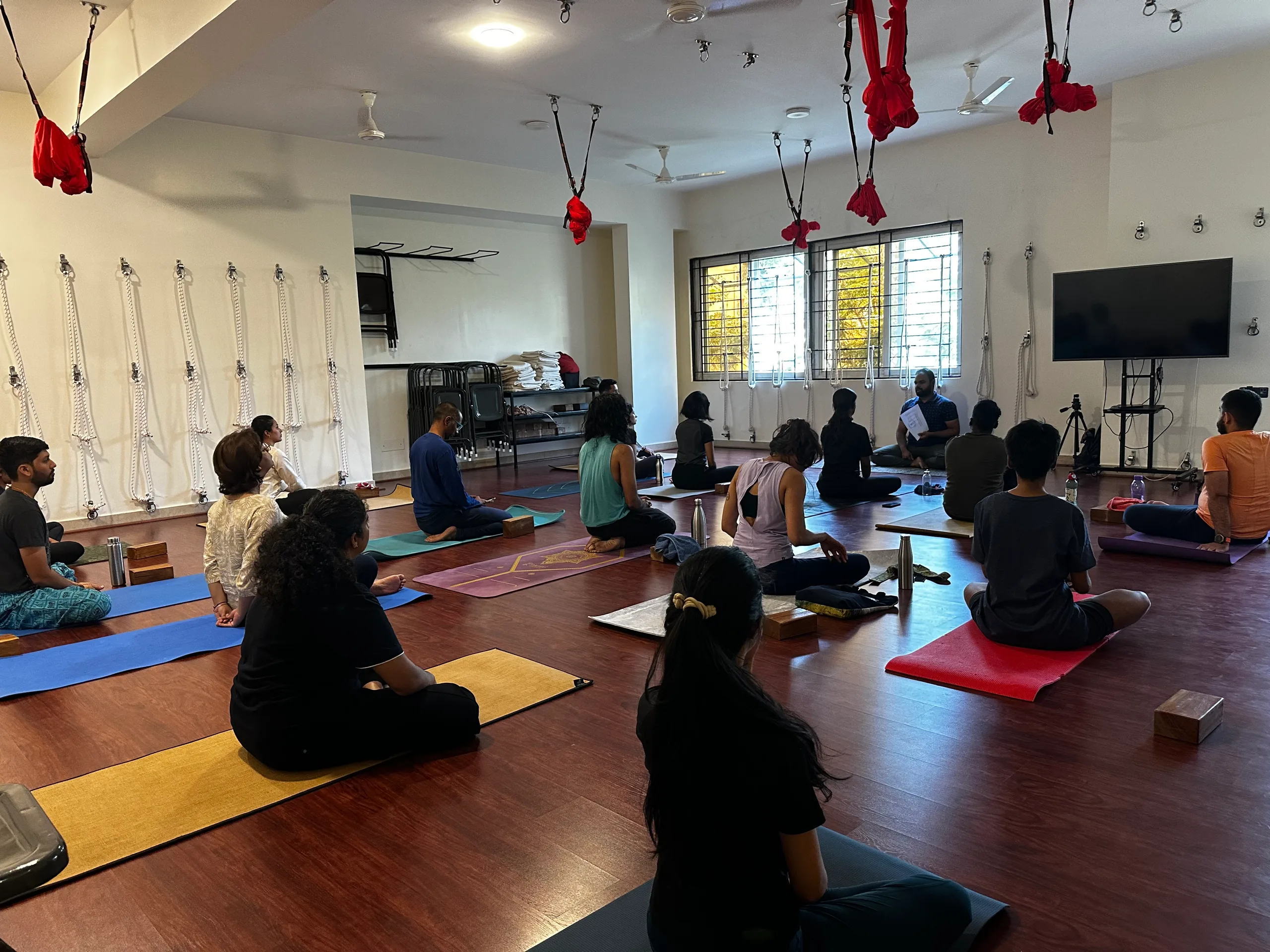 A wide view of a full studio: participants seated on coloured mats beneath ceiling slings, a rope wall along the left.