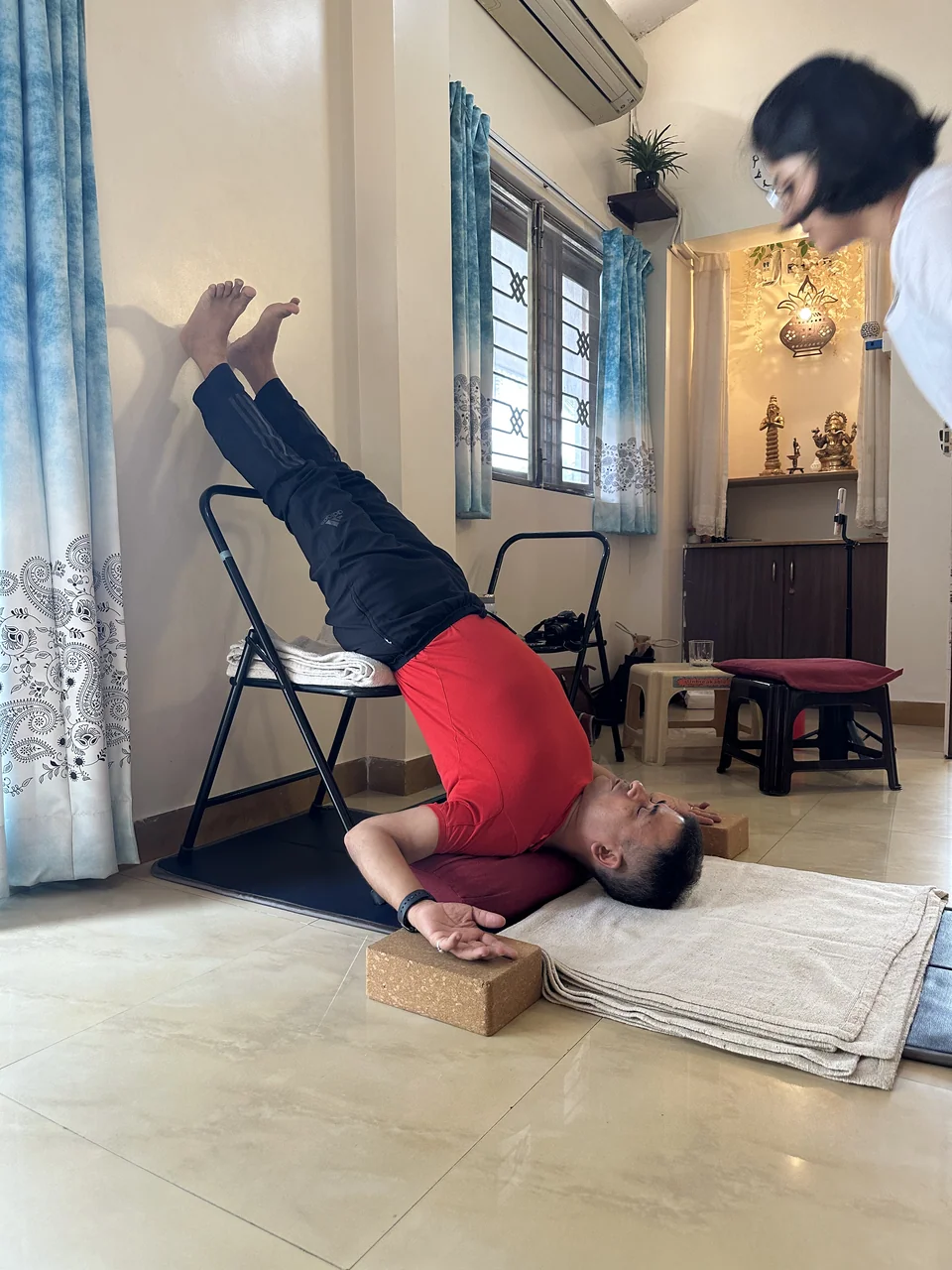 A man in a supported shoulderstand, his hips over a folding chair, his feet against the wall and his shoulders on a folded blanket between two blocks