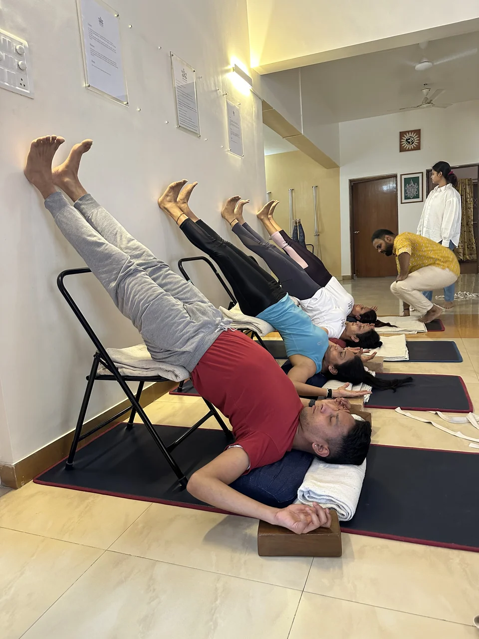 A man in supported shoulderstand in the foreground while a teacher bends over a student behind him