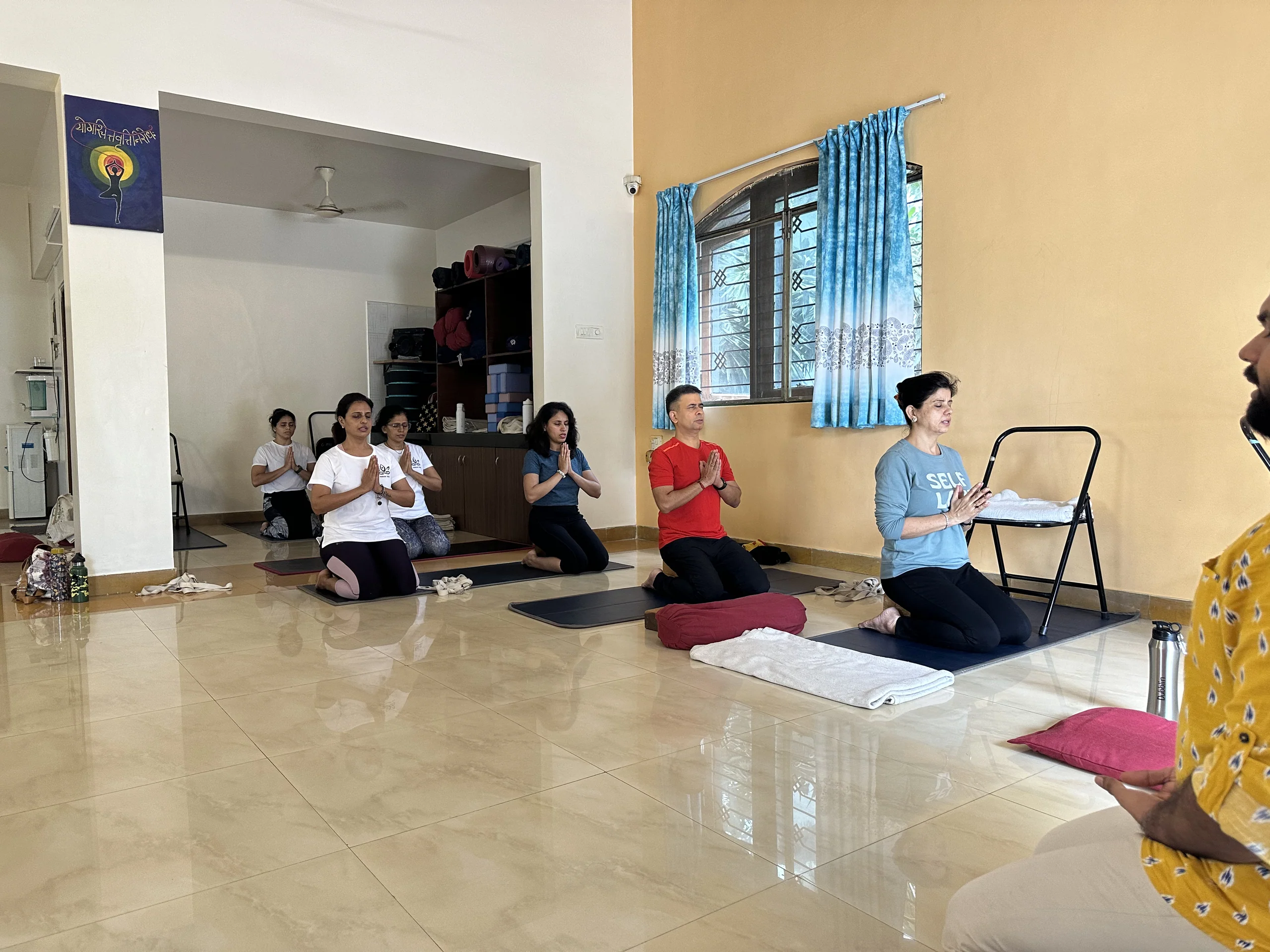 A group kneeling on mats with palms joined and eyes closed, in a room with a yellow wall and blue curtains