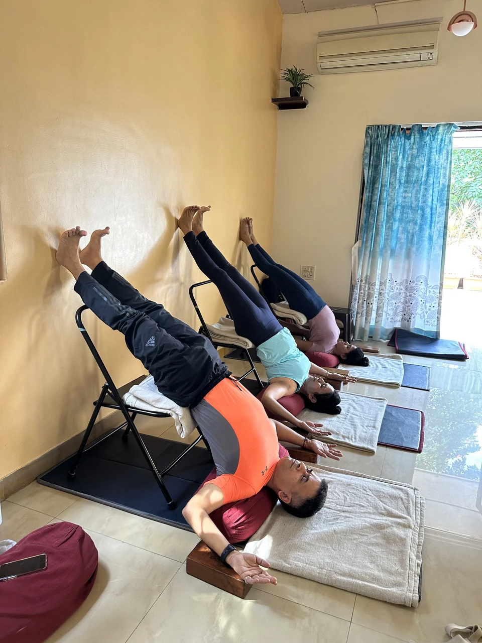 Three people in supported shoulderstand against a yellow wall with daylight from an open window