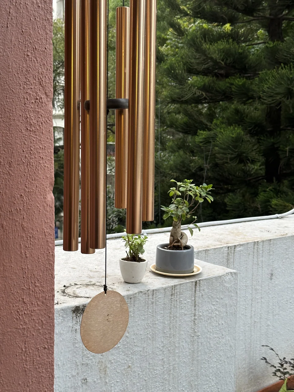 Brass wind chimes and a wooden striker disc hanging above a white parapet with trees behind
