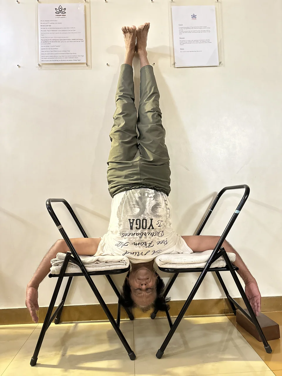 A person in a supported headstand between two chairs with arms spread along the seats