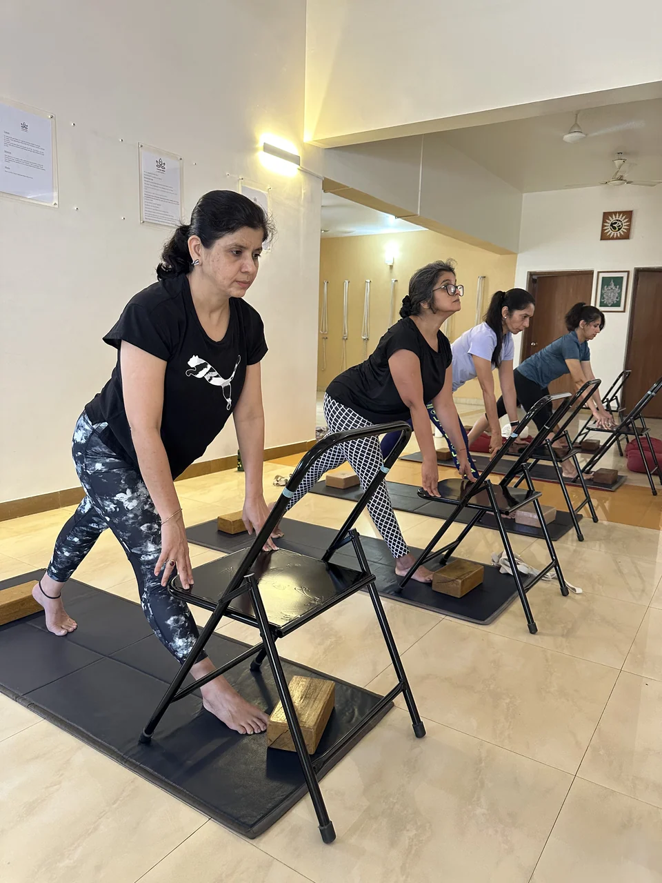 Four people in a line folding forward with their hands on the backs of folding chairs
