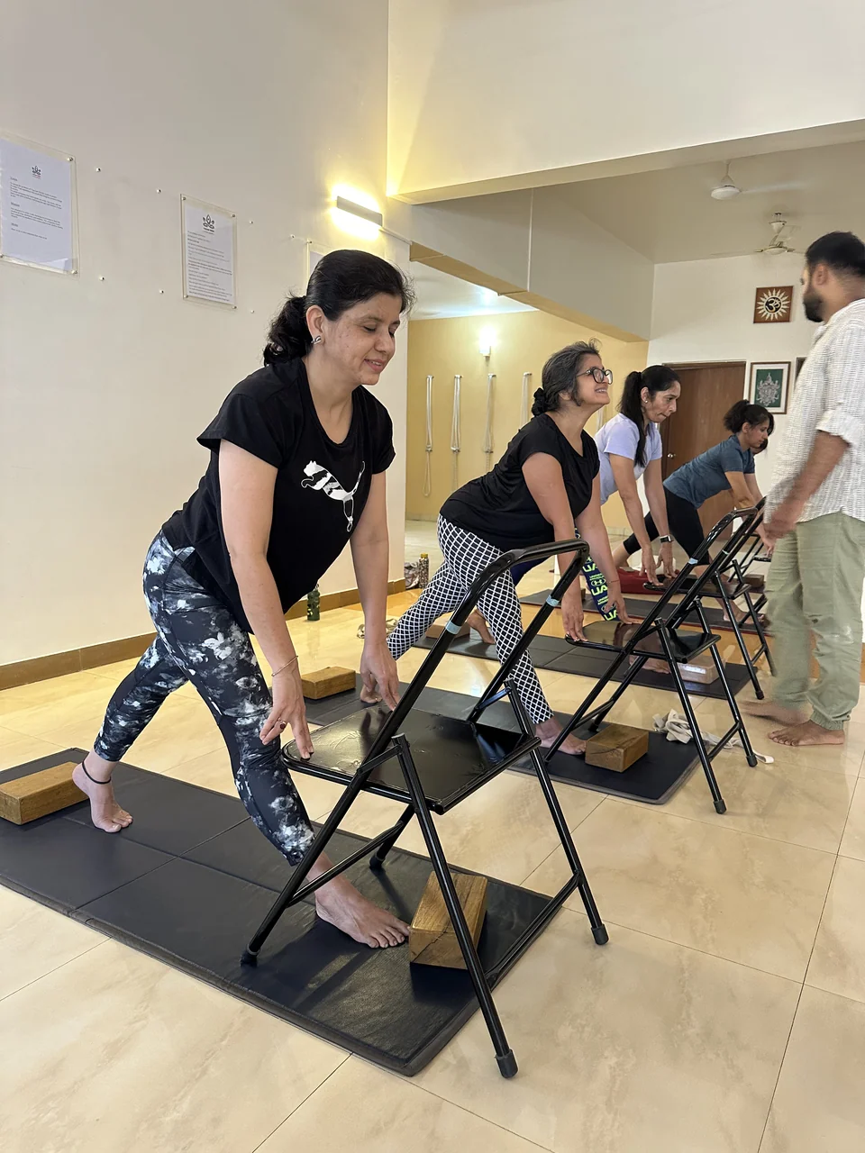 A line of people folding forward over chairs while a teacher adjusts the hands of one at the far end