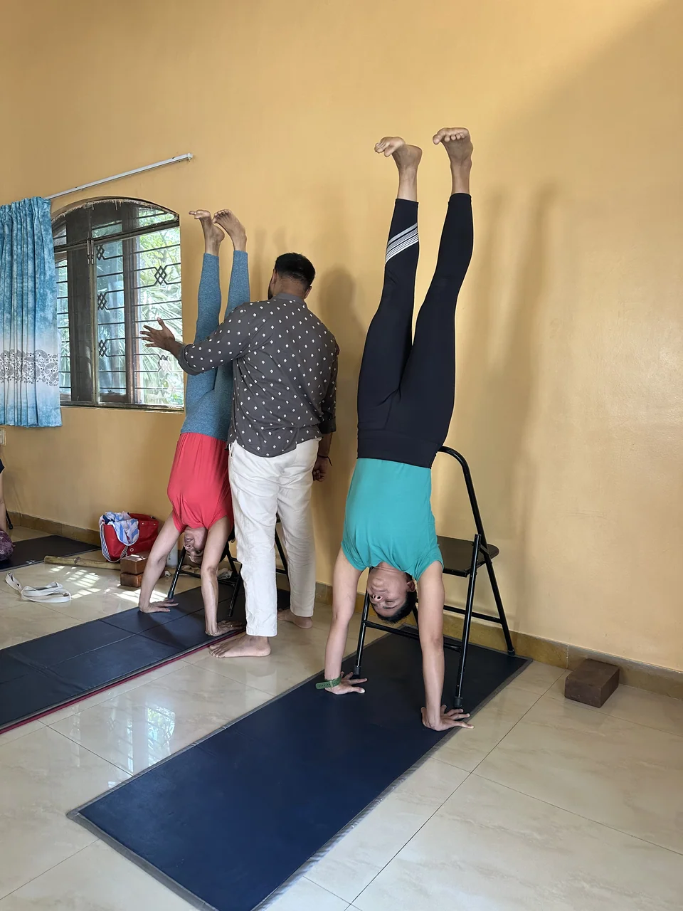 A person holding a handstand against a yellow wall while a man steadies another person's legs