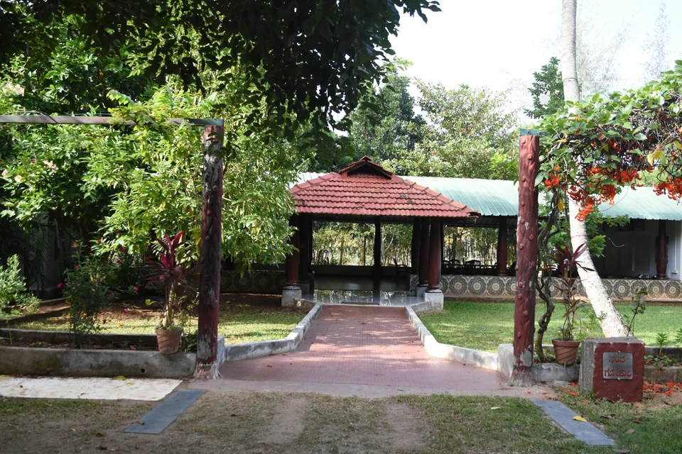 An open-sided pavilion with a red tiled roof at the end of a red paved approach, framed by flowering creeper