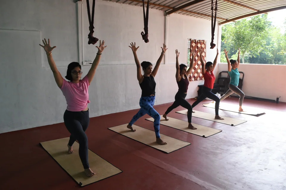 Five practitioners in warrior one with arms raised on mats across a red floor