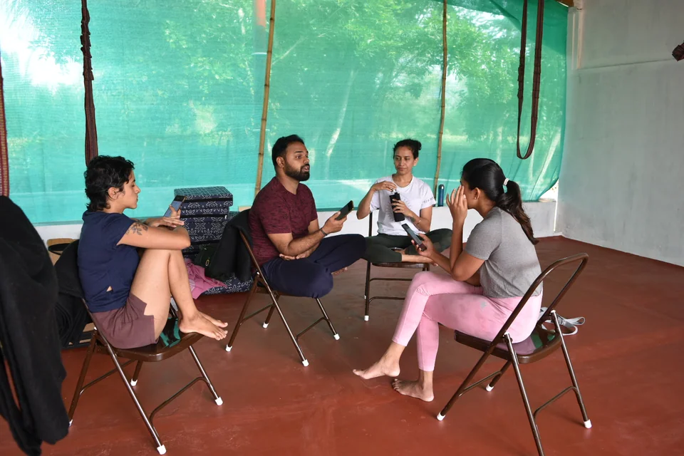 Five people sitting on chairs and the floor in a loose circle talking, green netting behind them