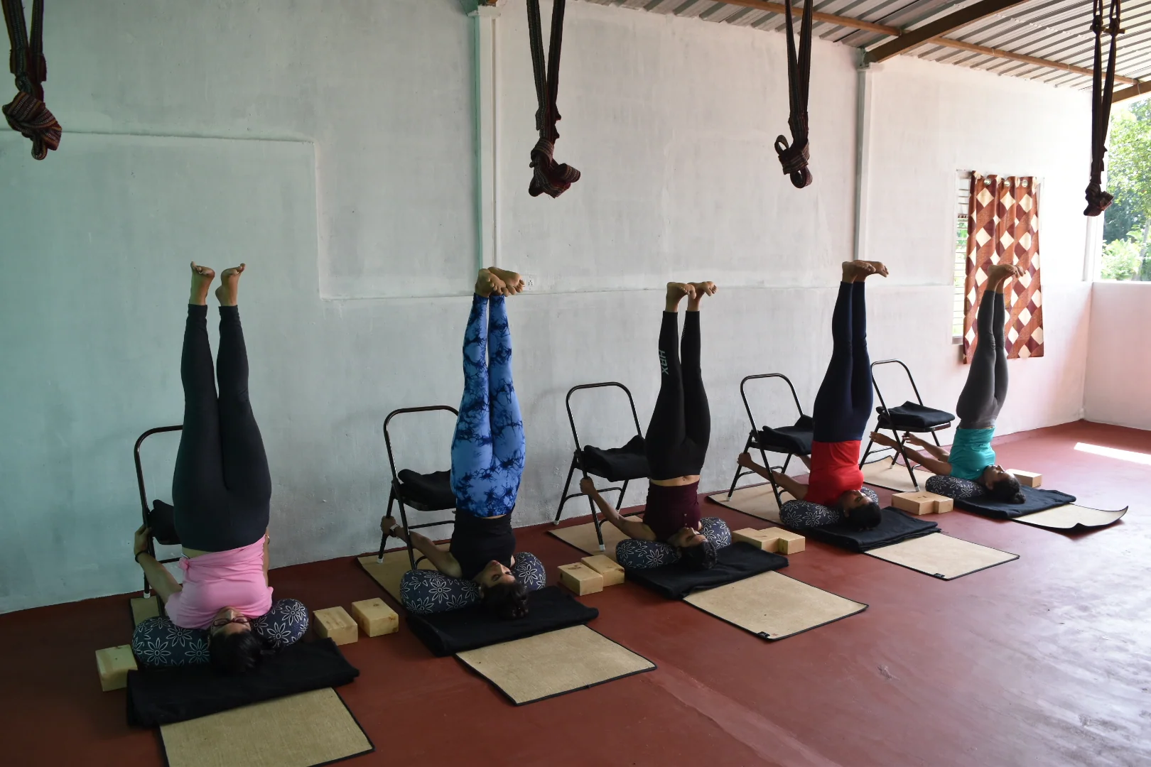 Four people in supported headstand over folding chairs in a row along a white wall