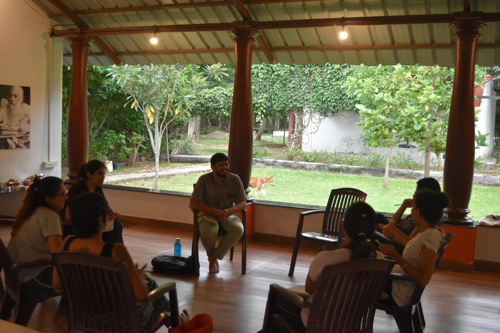 A group seated in a circle on chairs inside an open-sided pavilion looking out onto a lawn and trees