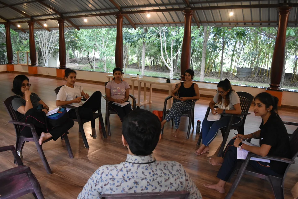 A discussion circle in an open pavilion with several people writing in notebooks as one speaks