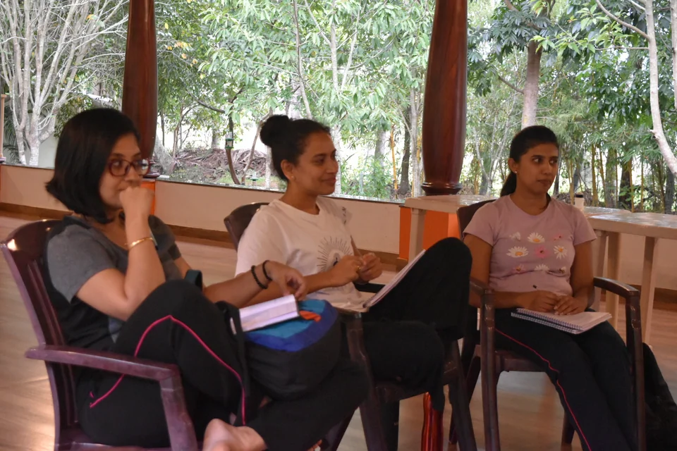 Three women seated on chairs listening, one resting her chin on her hand, another holding a notebook