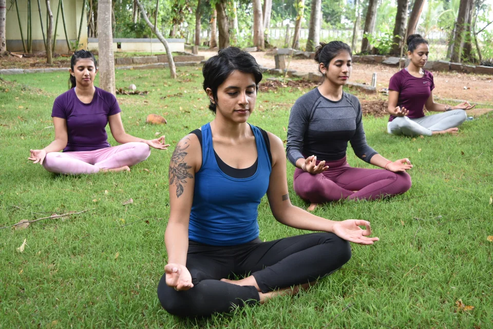 A woman sitting cross-legged on grass with her eyes closed and hands on her knees, three others seated behind