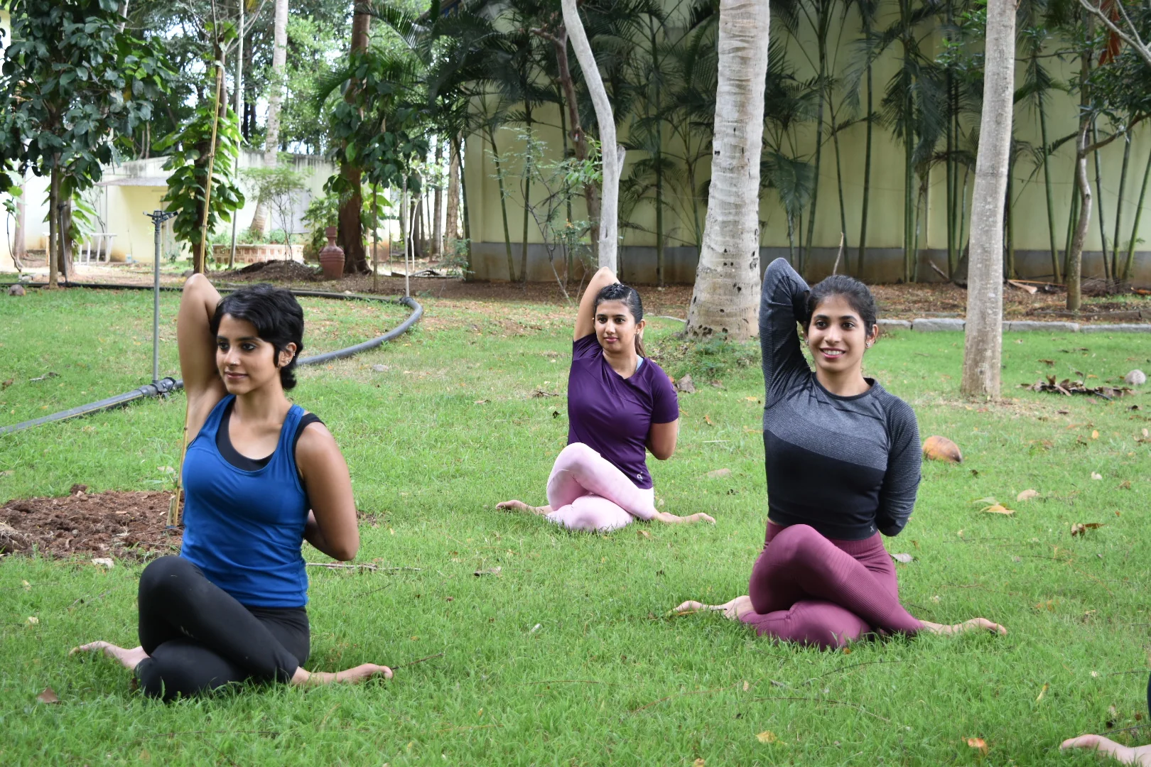 Three women sitting cross-legged on grass with one arm raised and bent over the head
