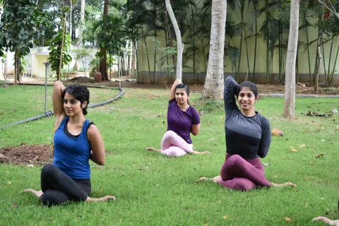 Three women sitting cross-legged on grass with one arm raised and bent over the head