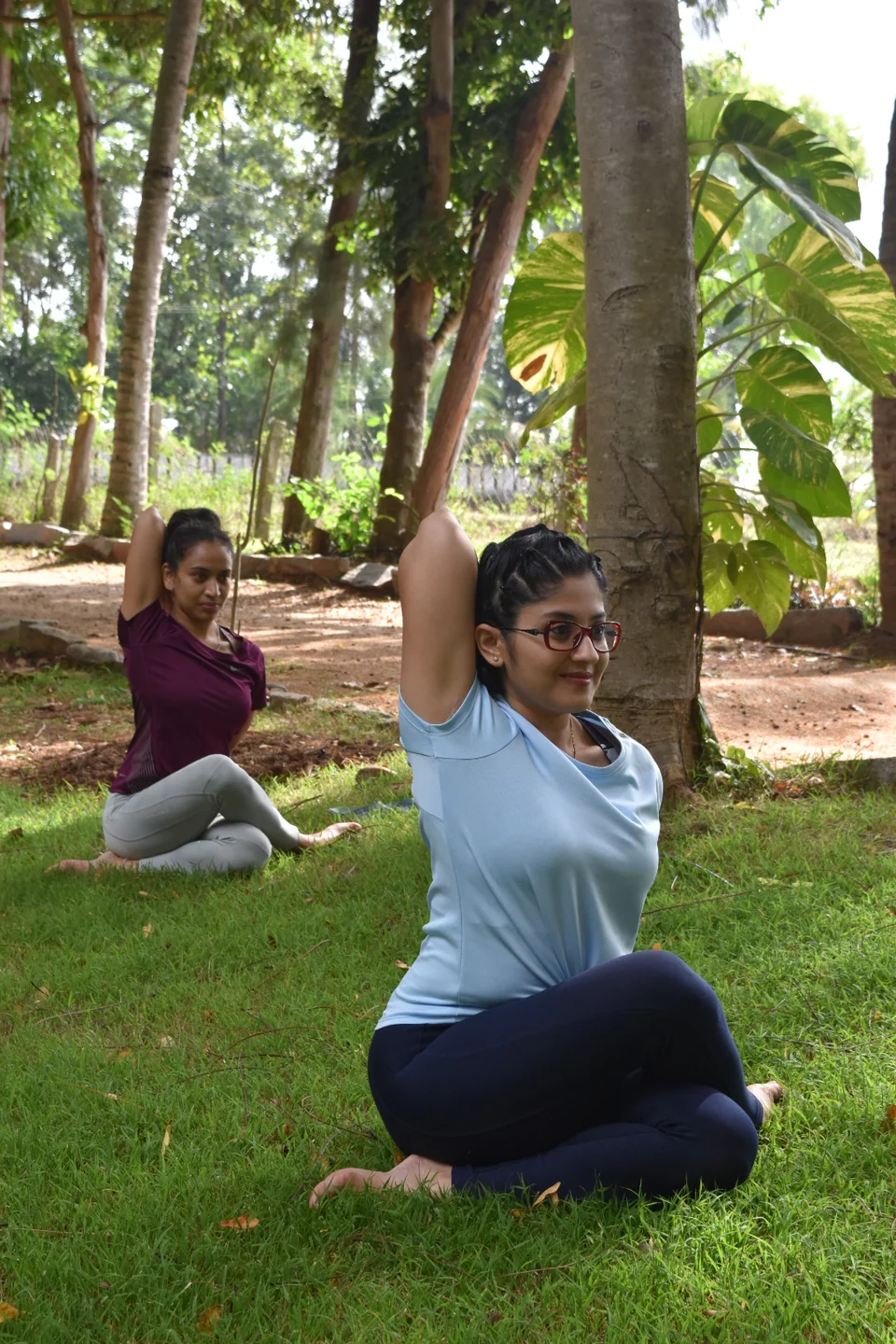 A woman sitting cross-legged on grass with both arms raised overhead, another practitioner behind