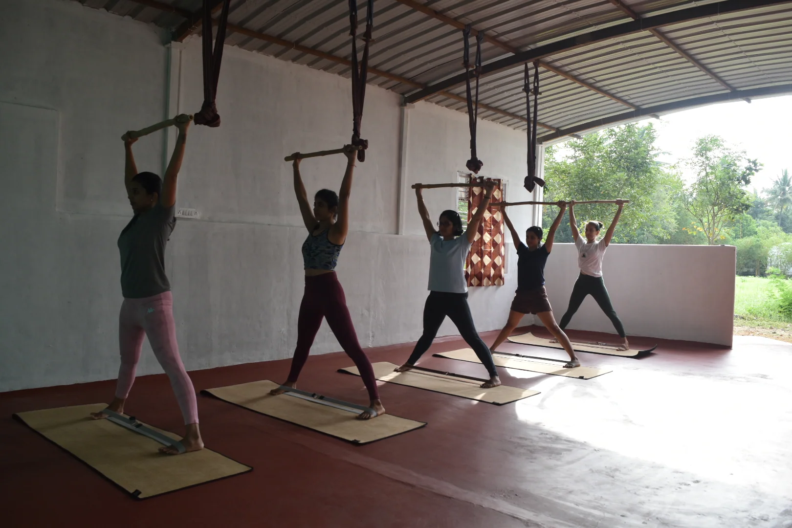 Five practitioners standing with arms raised overhead in an open-sided hall, low light across the floor