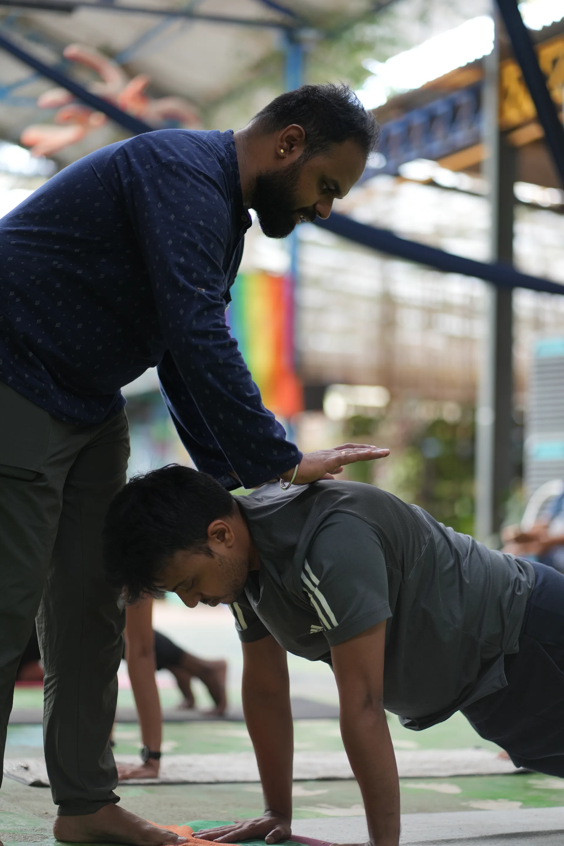A teacher bends over a student holding a plank, one hand just above the student's back to correct the line of the pose