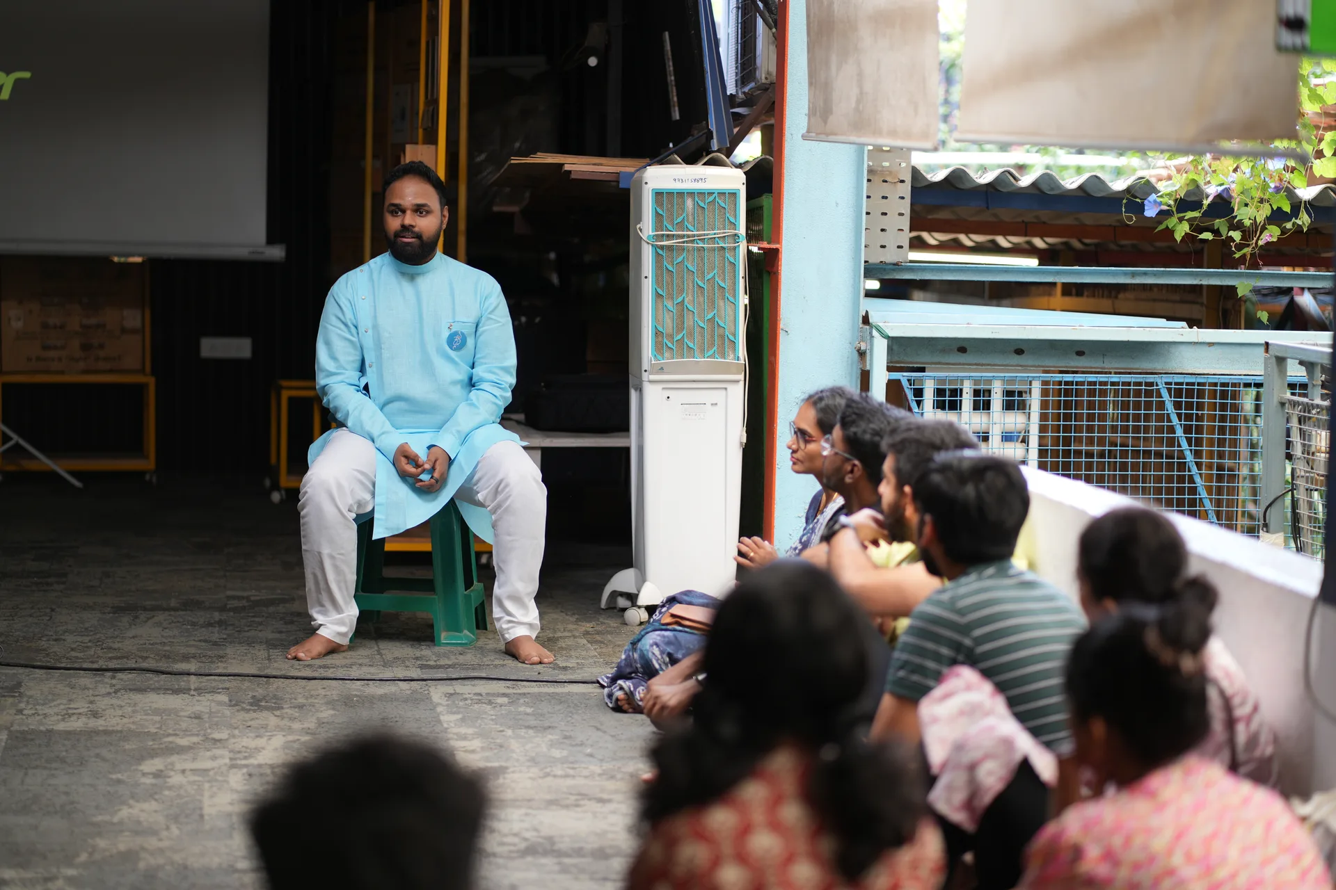 A man in a light blue kurta sits on a low stool speaking, with listeners seated on the floor before him