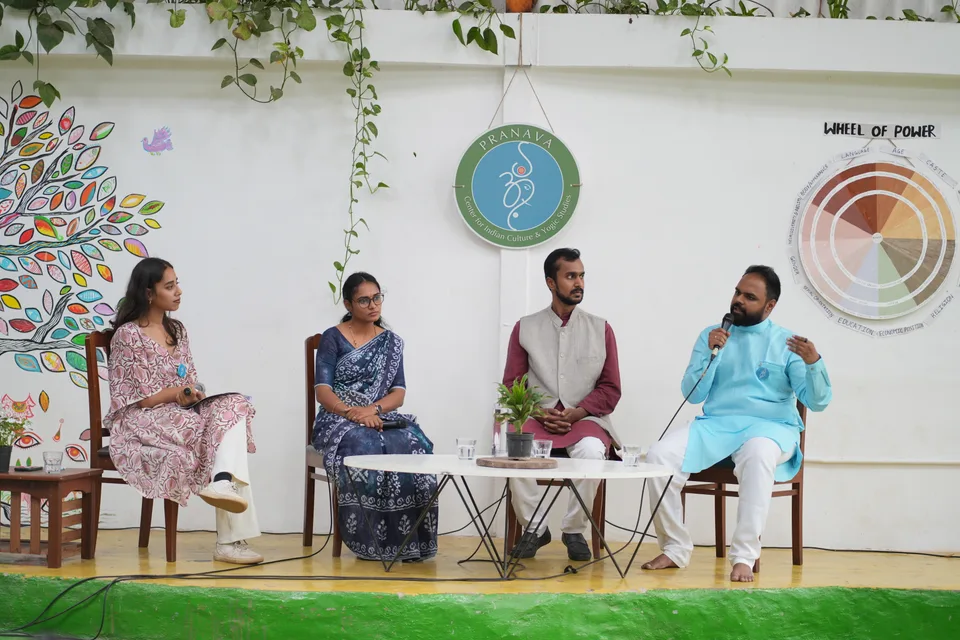 Four people seated on wooden chairs on a low stage, the man at the right speaking into a handheld microphone, a Pranava medallion on the wall behind them.