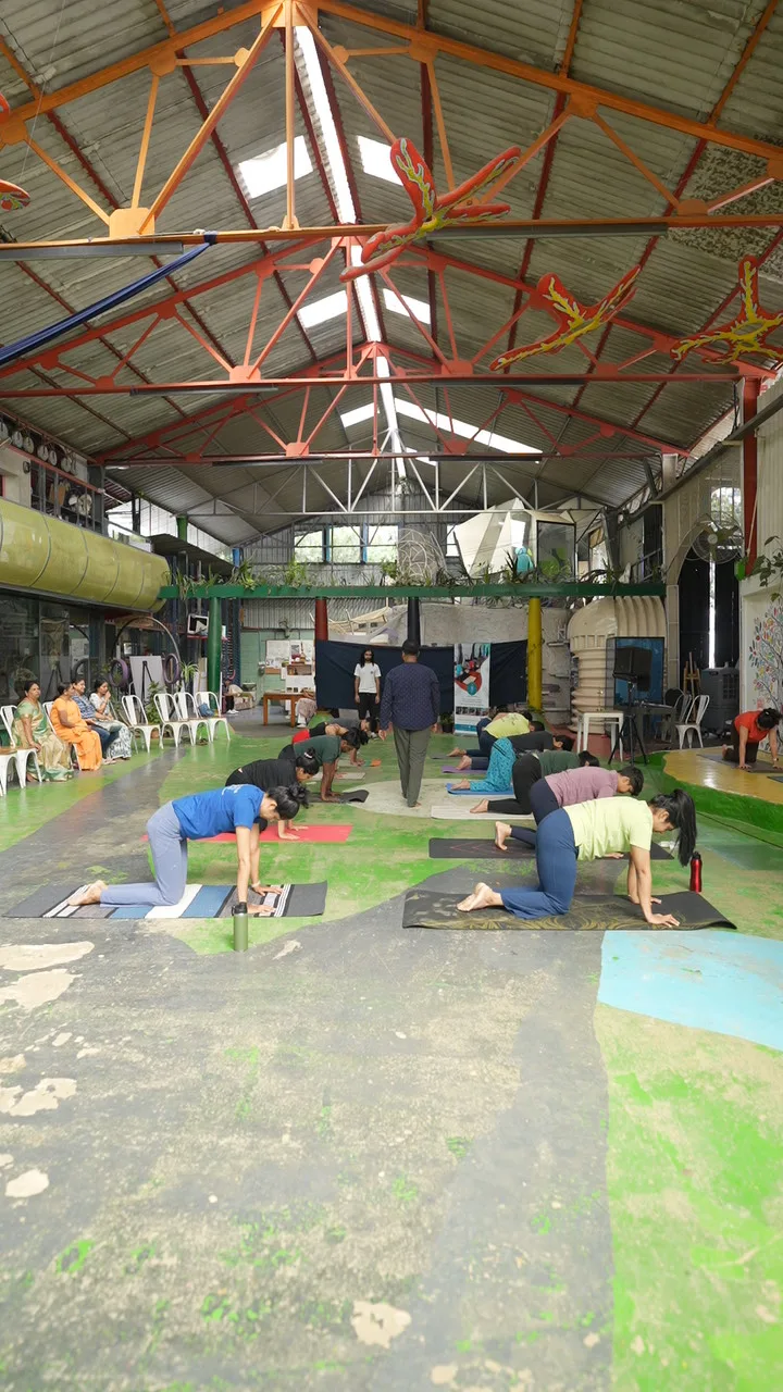A covered hall with red roof trusses and skylights above a line of practitioners bending over mats.